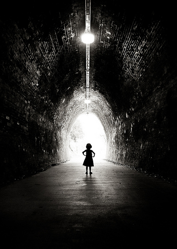 Low key black and white photo of girl in tunnel silhouetted against light at the end of the tunnel