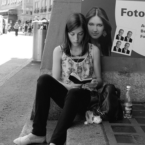 Street photo of girl reading with a poster behind looking over her shoulder, photographed using a large sensor compact camera