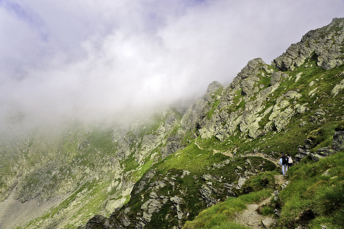 Two hikers on a narrow mountain path, clouds rolling over the edge of the mountain in front of them