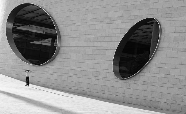 Photograph of a skateboarder skating past a building with giant circular windows. Taken with a 50mm prime lens.