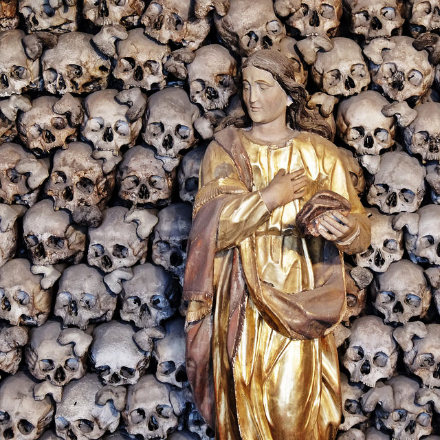 Photo of a statue with a wall of skulls behind it. Taken with a Compact System Camera then edited in Snapseed for iOS.