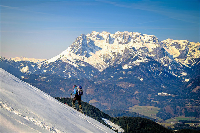 Walker with skies ascending a snowy mountain, with more snow capped mountains in the background. Captured using a kit lens.